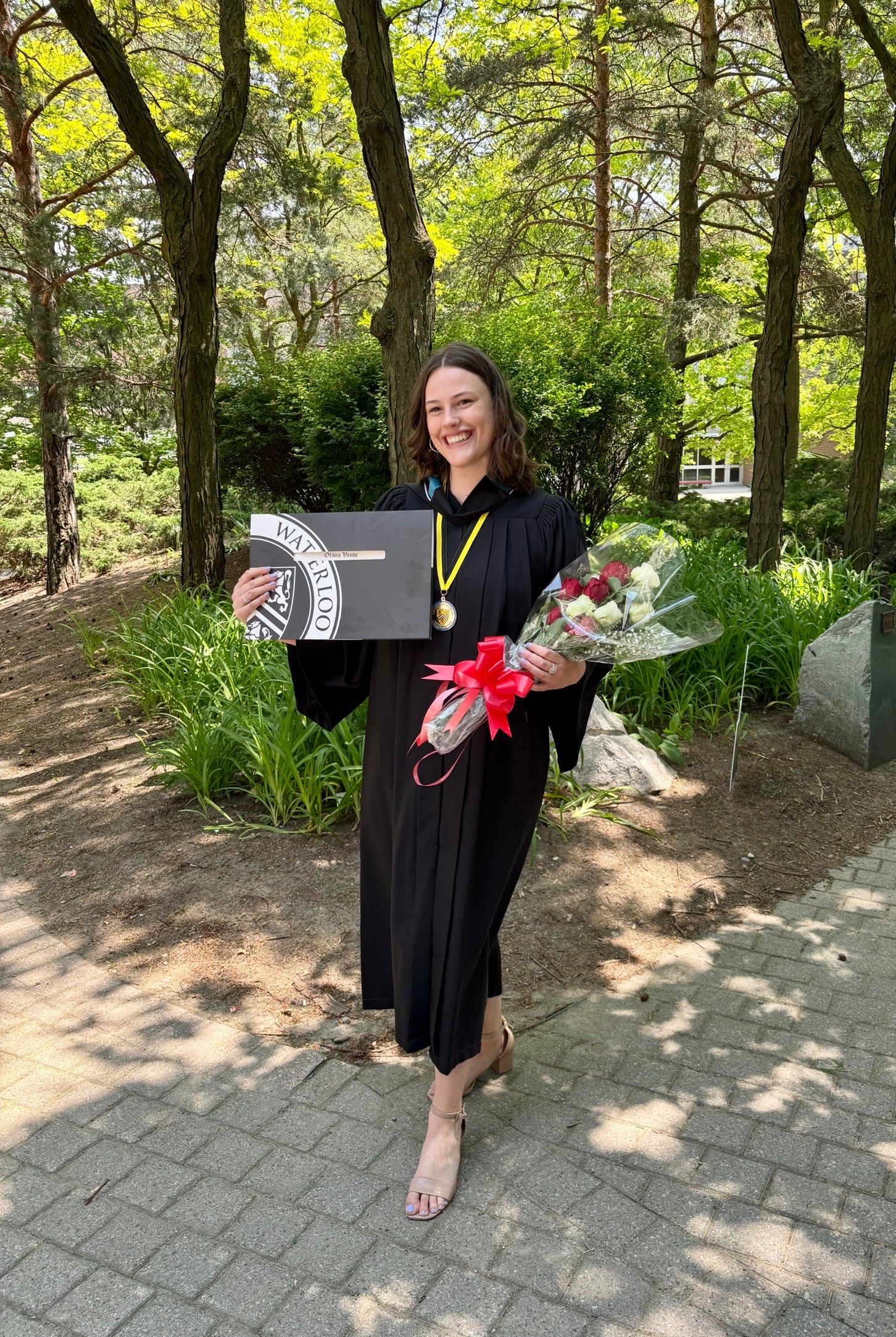 Olivia on her graduation day in cap and gown holding a degree and flowers outdoors.